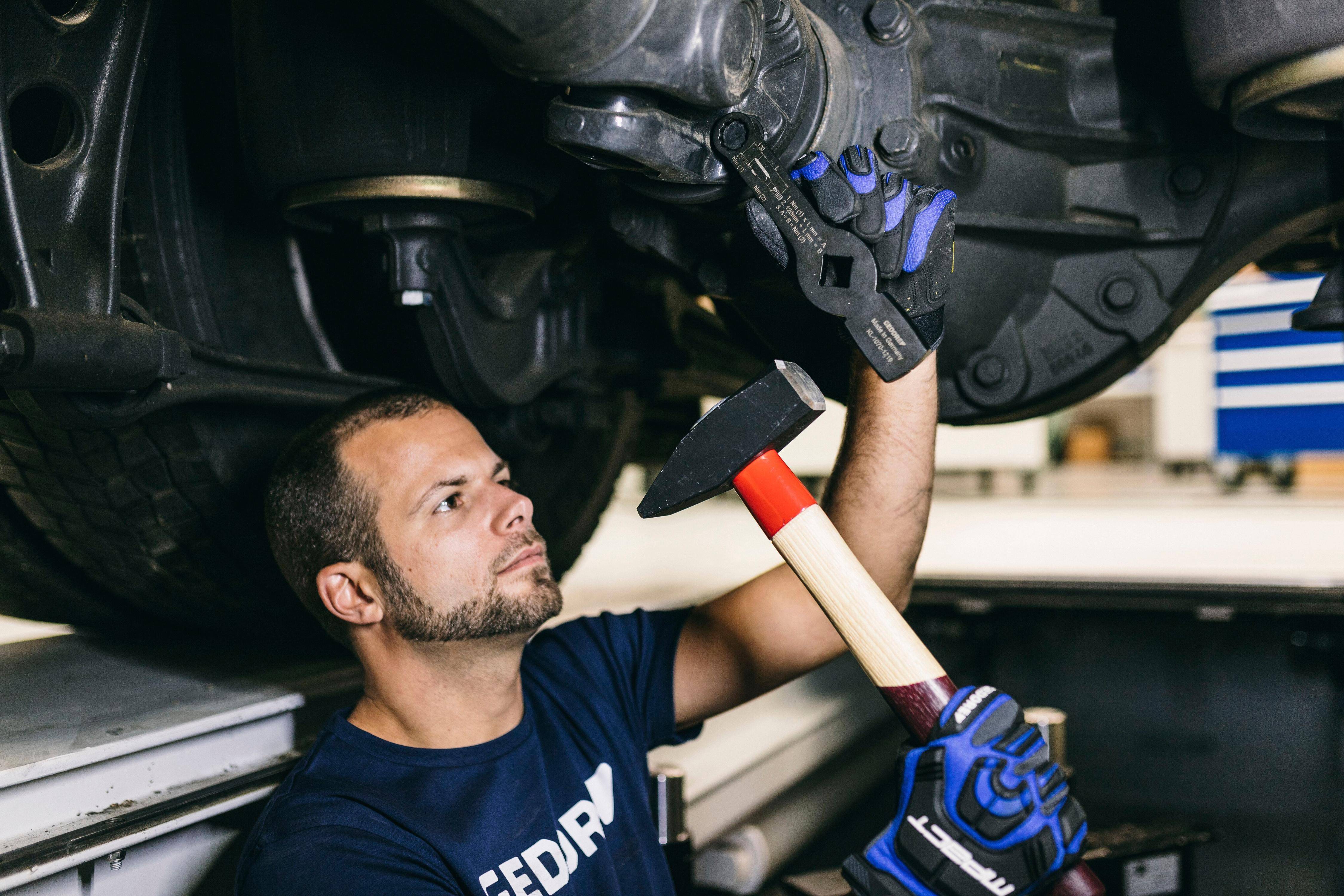 Ein Mechaniker zieht eine Schraube unter einem Auto mit einem großen Schraubenschlüssel und einem Hammer fest, trägt blaue Handschuhe und konzentriert sich auf die Aufgabe.