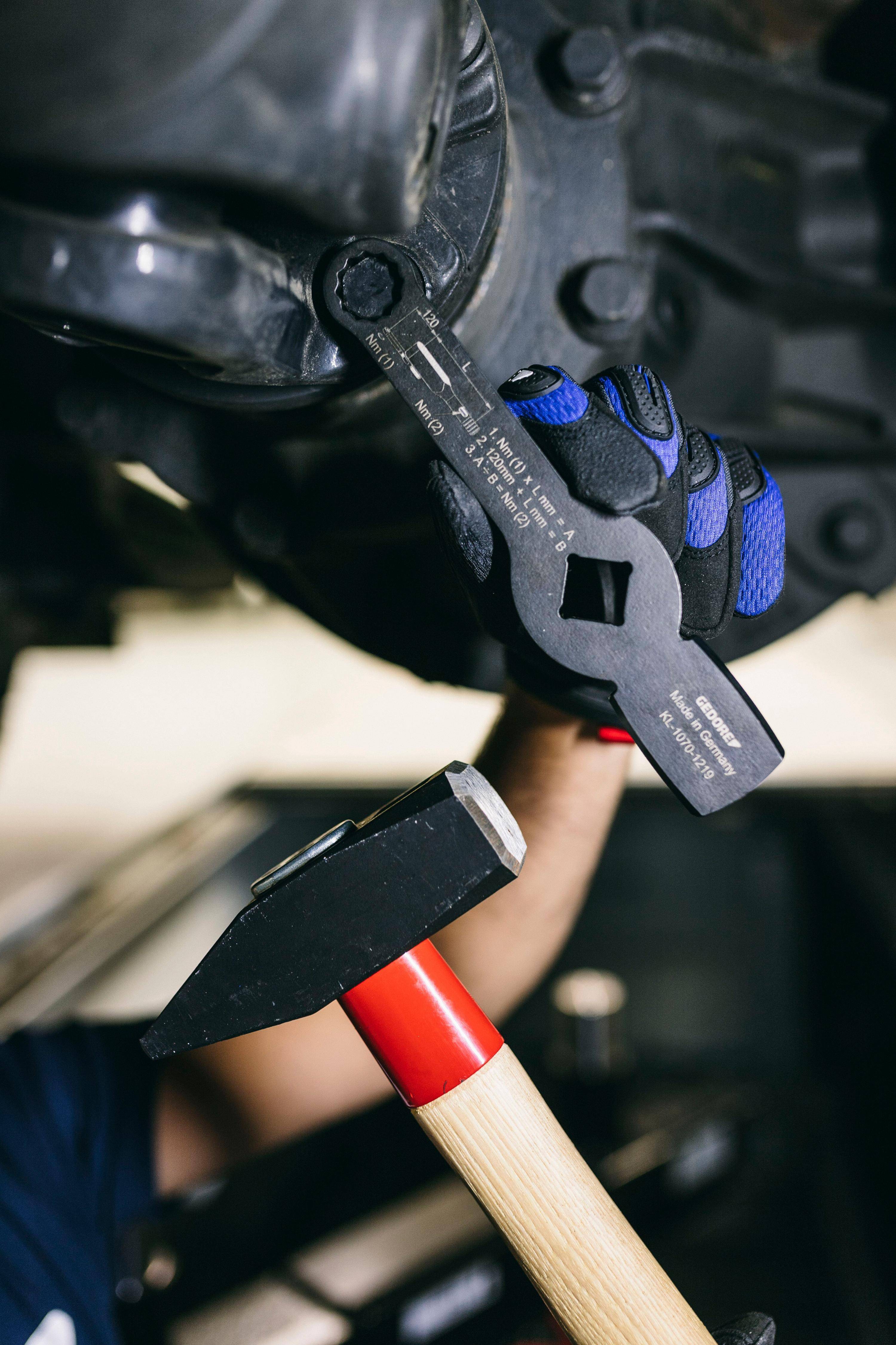 Eine Person benutzt einen schwarzen verstellbaren Schraubenschlüssel an einem Autoteil. Eine behandschuhte Hand greift den Schraubenschlüssel, und ein Hammer ist darunter sichtbar.