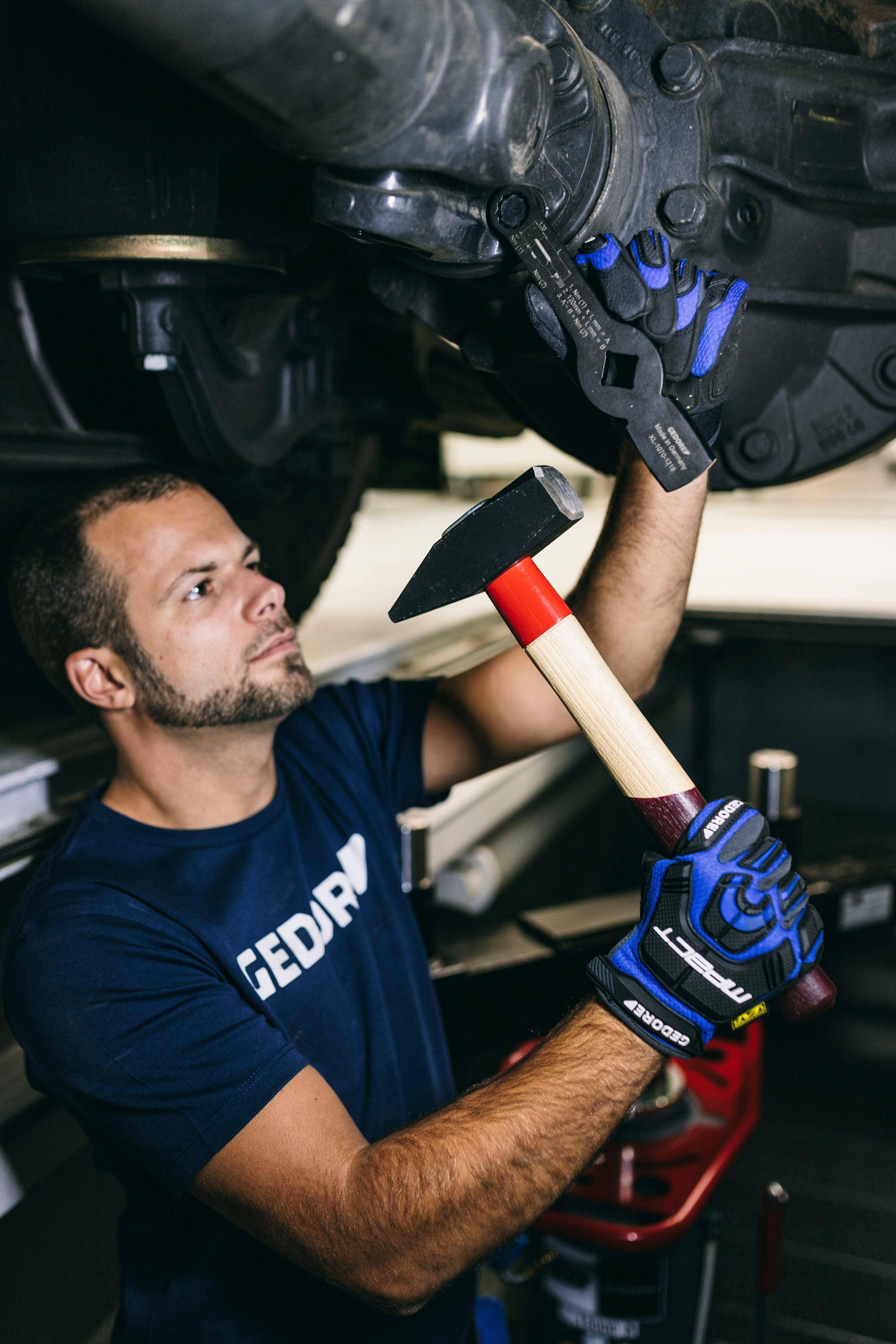 Ein Mechaniker in Handschuhen arbeitet unter einem Fahrzeug und nutzt ein Werkzeug, um ein Bauteil einzustellen. Der Fokus liegt auf der Werkzeugnutzung und der Einstellungsaufgabe.