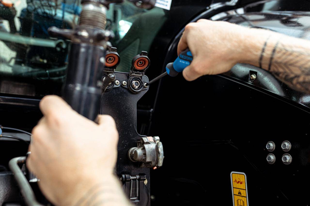 Eine Person benutzt einen Schraubenzieher, um ein Metallbauteil an einer Maschine einzustellen und konzentriert sich dabei auf Präzision. Verschiedene Schrauben und Teile sind sichtbar.