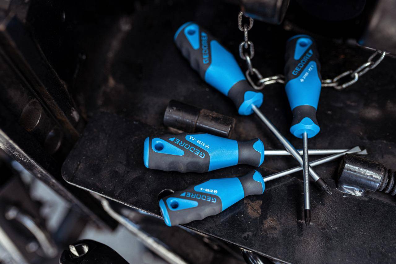 Vier blau-schwarze Schraubendreher in verschiedenen Größen liegen verstreut auf einer dunklen Oberfläche, wobei im Hintergrund ein Maschinenteil sichtbar ist, was auf eine mechanische Arbeitsumgebung hindeutet.