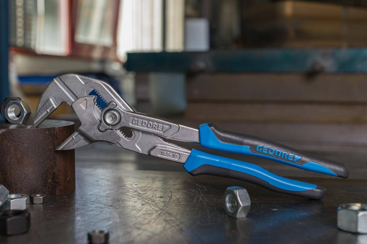 Zange mit blauen Griffen, die einen rostigen Metallzylinder auf einer Werkbank ergreift; eine Mutter liegt in der Nähe.