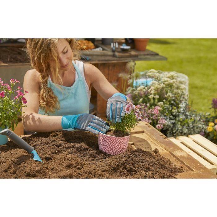 Frau in Handschuhen pflanzt eine kleine Blütenpflanze in einen rosa Topf in ein Gartenbeet, mit Erde und Gartengeräten in der Nähe.