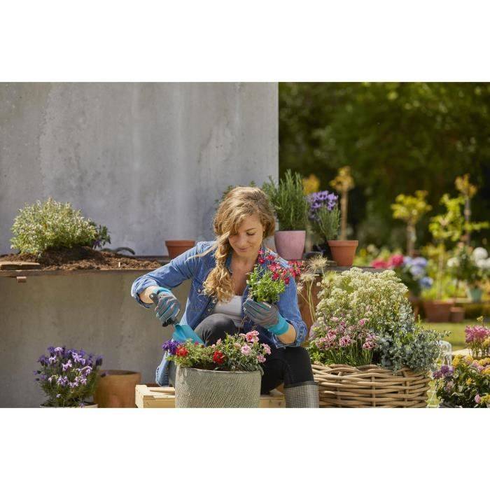 Frau arbeitet im Freien im Garten und pflanzt Blumen in einen Behälter neben Körben und Topfpflanzen in einem sonnigen Hof.