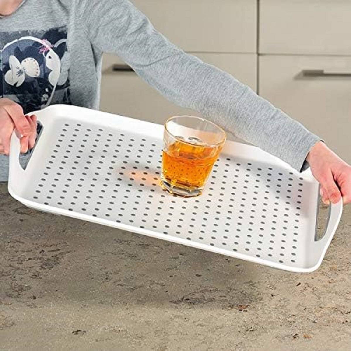 Person, die ein weißes durchlöchertes Serviertablett mit einem Glas eines bernsteinfarbenen Getränks darauf hält.