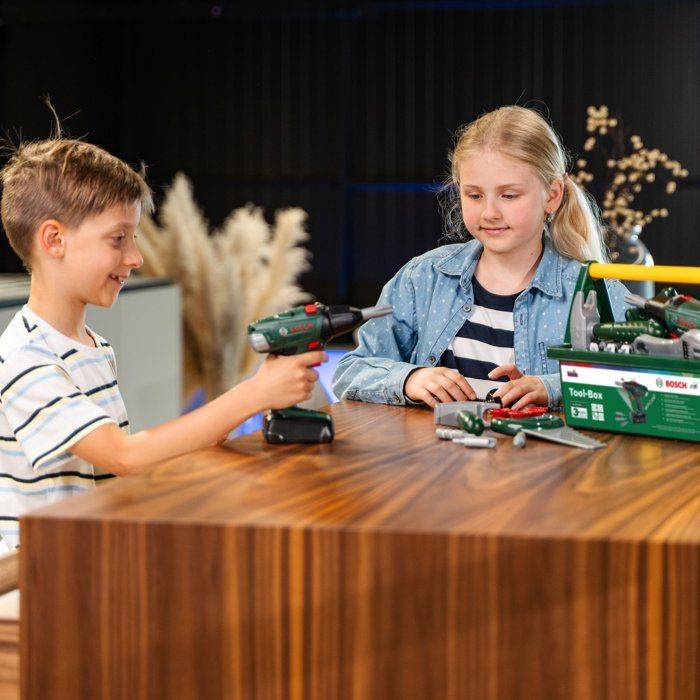 Junge bietet einem Mädchen an einem Tisch mit Spielzeugwerkzeugen und einer Bosch-Werkzeugset-Box einen Spielzeugbohrer an.