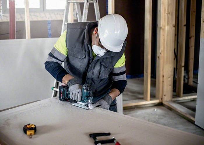 Ein Bauarbeiter mit Schutzausrüstung arbeitet mit Elektrowerkzeugen an einer Werkbank in einem teilweise fertiggestellten Raum.