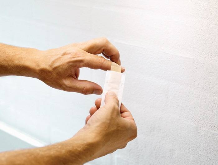 Eine Person, die einen Verband an ihrem Finger anlegt, was Selbstfürsorge oder erste Hilfe andeutet, vor einem einfarbigen Wandhintergrund.