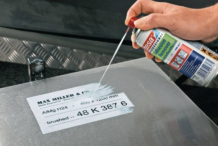 Eine Hand sprüht ein Reinigungsmittel auf metallische Klebereste neben einem Etikett mit der Aufschrift 