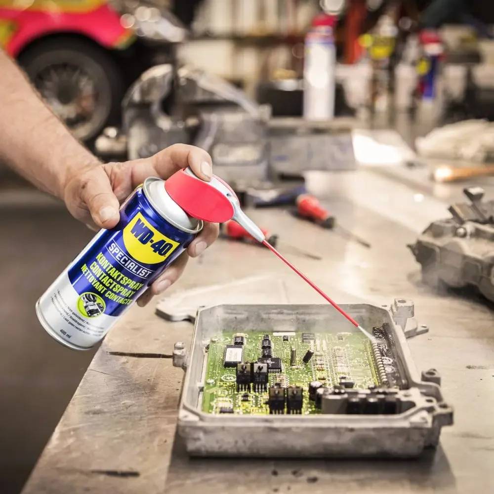 Eine Person sprüht WD-40 auf eine freiliegende elektronische Leiterplatte in einem Metallgehäuse auf einer Werkbank.