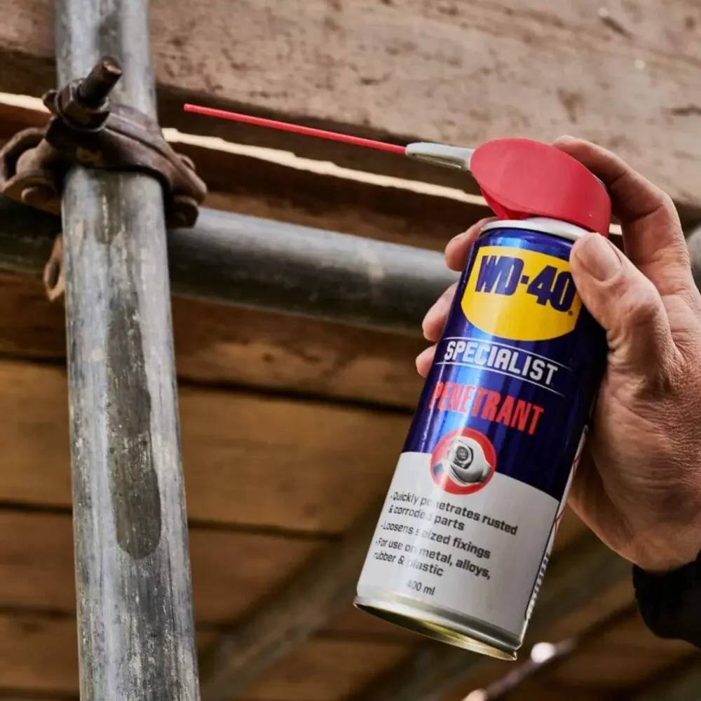 Eine Hand hält eine WD-40 Specialist Penetrant-Sprühdose mit rotem Strohhalm, der auf eine rostige Metallgerüstklammer und ein Rohr abzielt.