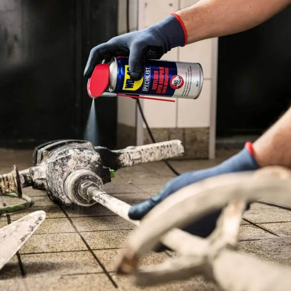 Eine behandschuhte Person sprüht Schmiermittel aus einer WD-40-Dose auf ein schmutziges mechanisches Gelenk oder Wellenverbindung auf dem Boden.