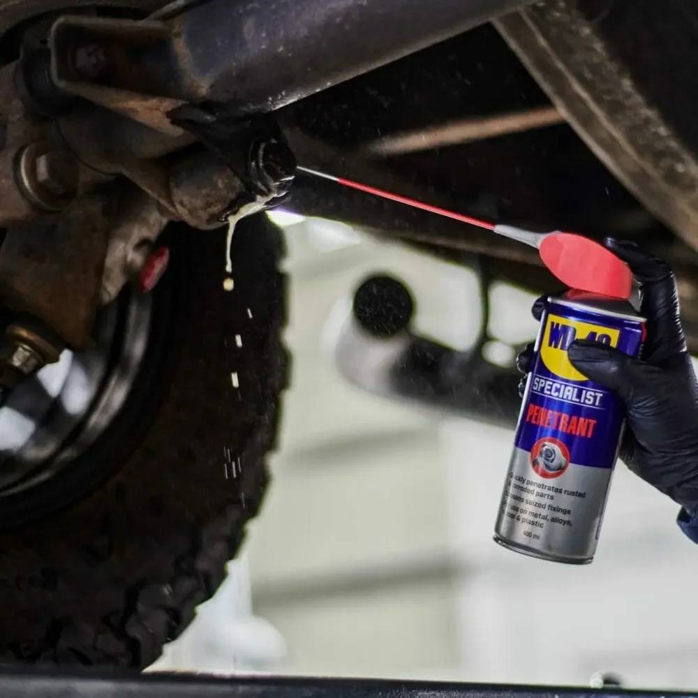 Hand mit Handschuh sprüht WD-40 Specialist Penetrant auf die Unterseite eines Fahrzeugs, um ein verrostetes Teil zu lockern, während Tropfen darunter herunterfallen.