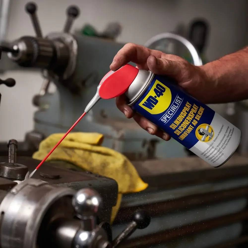Hand mit WD-40 Specialist Spray, um ein Metallmaschinenteil in einer Werkstatt zu schmieren.