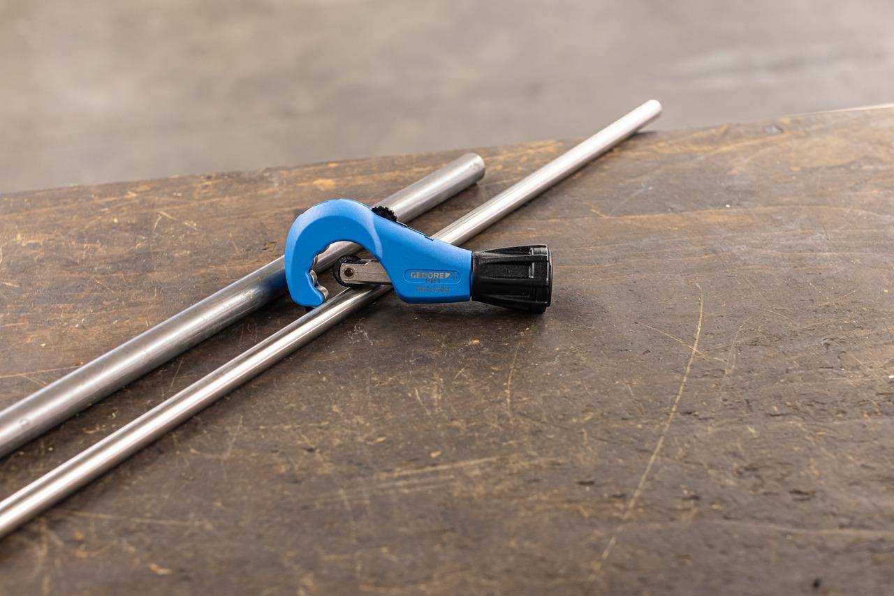 Ein manueller Rohrabschneider mit blauem Griff und schwarzem Handgriff ist auf einer Holzoberfläche positioniert, neben zwei Metallrohren, die zum Schneiden bereit sind.
