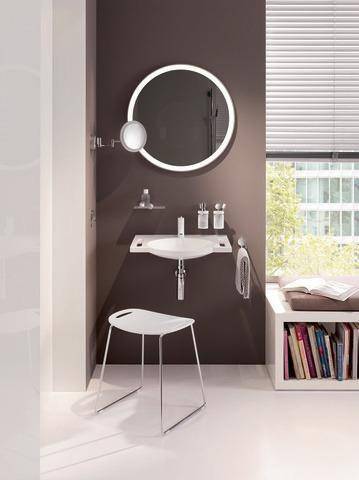 Ein modernes Badezimmer zeigt einen runden, hinterleuchteten Spiegel über einem weißen Waschbecken. Daneben steht ein weißer Stuhl, und Jalousien lassen natürliches Licht herein.