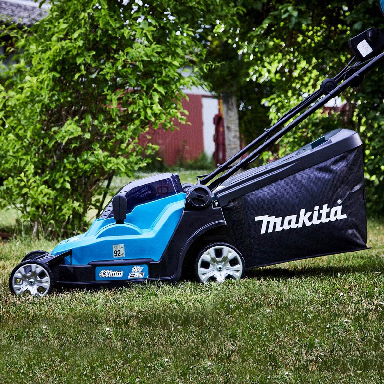 Ein blauer Makita-Rasenmäher steht auf einem grasbedeckten Rasen, umgeben von Grünpflanzen. Sein Griff ist hochgeklappt und bereit zur Verwendung. Das Markenlogo ist gut sichtbar.