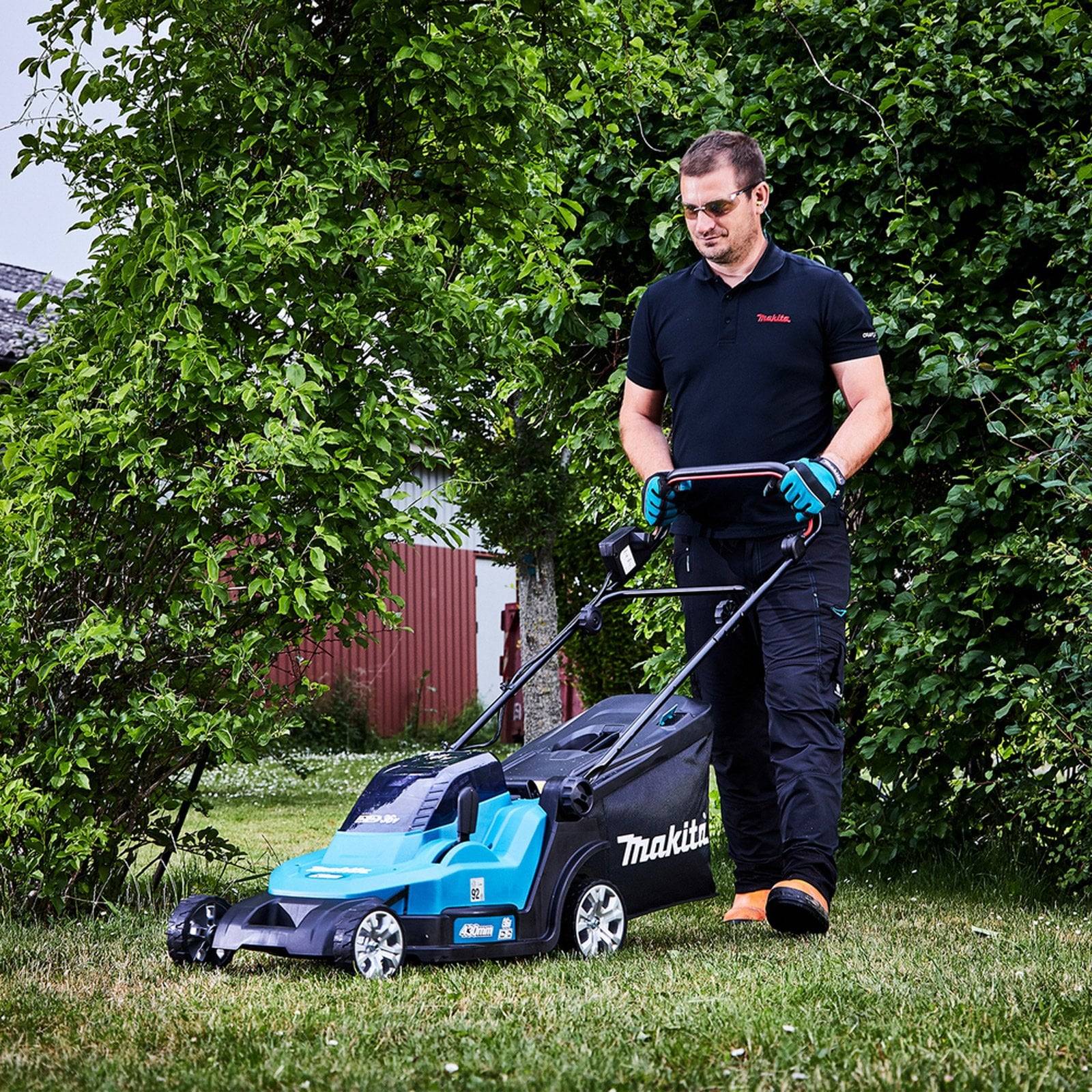 Eine Person schiebt einen blau-schwarzen Rasenmäher auf einem grasbewachsenen Grundstück. Sie trägt Handschuhe und Sonnenbrille und konzentriert sich aufs Rasenmähen. Im Hintergrund befinden sich Bäume und ein Gebäude.