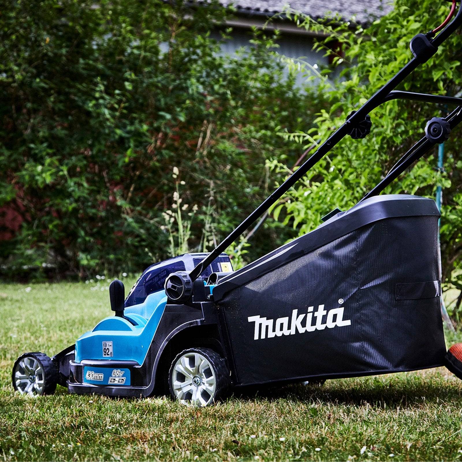 Eine blaue Makita-Rasenmähmaschine auf Gras, mit einem großen Grasfangbehälter. Im Hintergrund sind Büsche zu sehen.