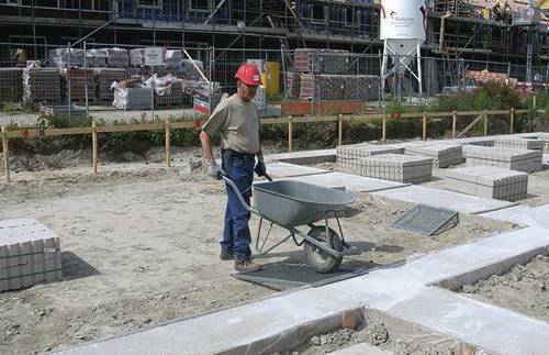 Ein Bauarbeiter mit rotem Schutzhelm schiebt eine Schubkarre über ein Betonfundament auf einer Baustelle.