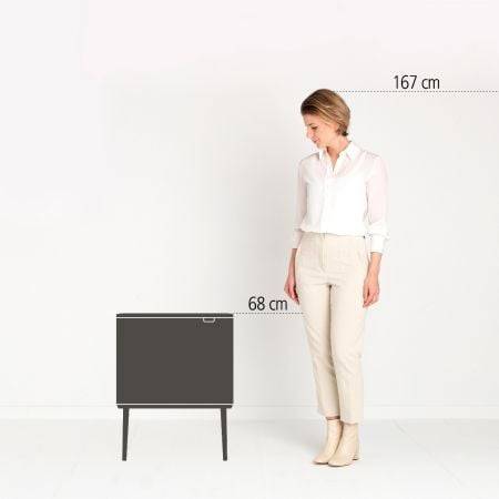 Eine Frau steht neben einem kleinen dunklen Schrank und zeigt seine Höhe im Vergleich zu ihrer Körpergröße von 167 cm; der Schrank ist 68 cm hoch.