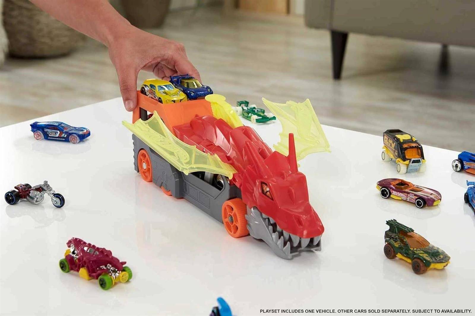Eine Hand platziert ein Spielzeugauto auf einem drachenförmigen Spielset mit Rampen, umgeben von mehreren kleinen Spielzeugautos auf einem weißen Boden.