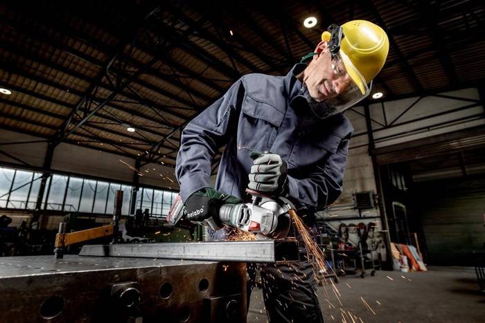 Arbeiter in einem gelben Schutzhelm verwendet einen Winkelschleiffer an einem Metallwerkstück und erzeugt Funken in einer Industriewerkstatt.