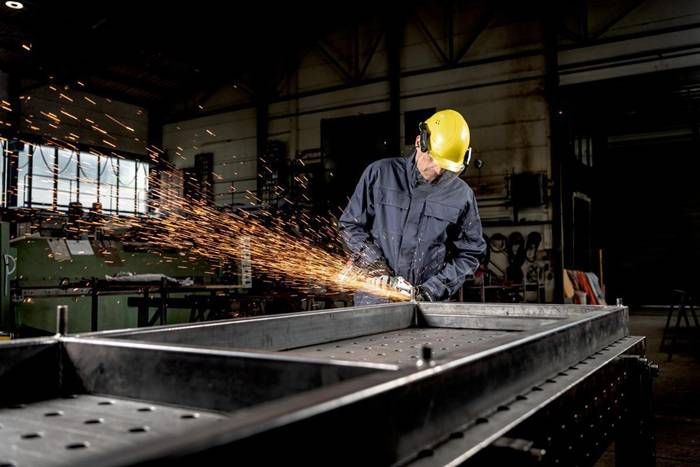 Arbeiter in gelbem Schutzhelm nutzt einen Winkelschleifer an einem Metallrahmen und wirft Funken in einer Industriewerkstatt auf.