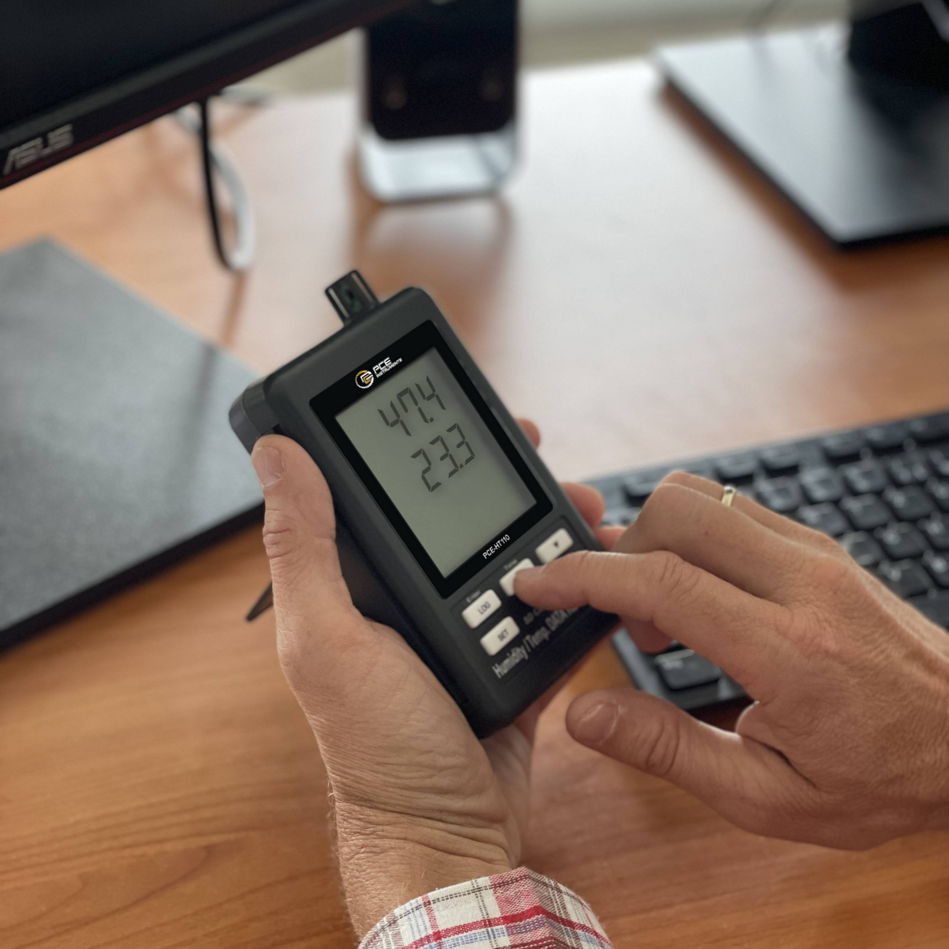 Eine Person hält ein digitales Gerät, auf dem '473,4' angezeigt wird, an einem Schreibtisch mit einem Computer und einer Tastatur. Der Fokus liegt auf der Anzeige des Geräts.