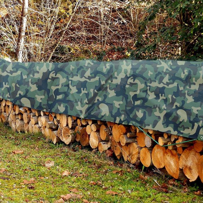 Aufgestapeltes Brennholz mit einer grünen Tarntplane bedeckt, im Freien auf Gras aufgestellt, umgeben von Bäumen und einem Zaun.