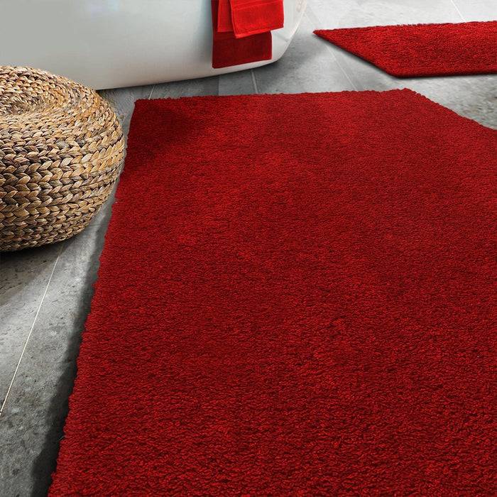 Ein roter Teppich auf einem grauen Fliesenboden neben einem Flechtkorb, mit einem Blick auf ein rotes Handtuch, das an einer weißen Badewanne hängt.