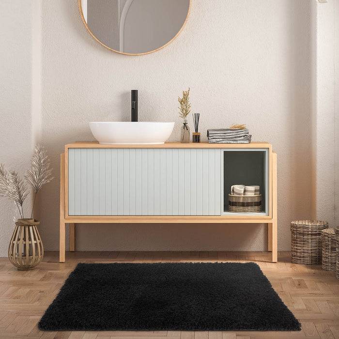 Ein minimalistisches Badezimmer mit einem runden Spiegel, einem Aufsatzbecken und einem Holzschrank auf einem Parkettboden mit einem schwarzen Teppich, einem Korb und einer Vase.