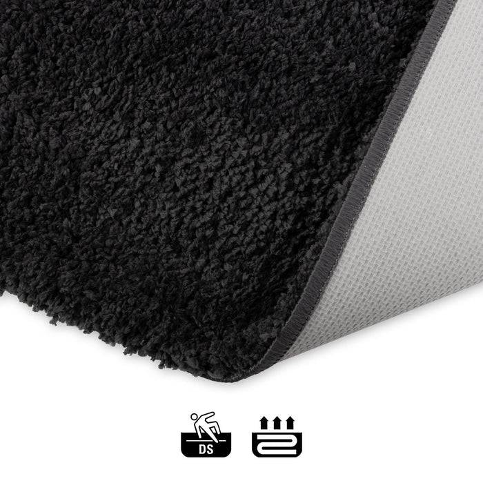 Eine Nahaufnahme einer schwarzen, strukturierten Ecke eines Teppichs mit einer hellgrauen Unterseite. Zwei Icons darunter zeigen an, dass er rutschfest und für Fußbodenheizung geeignet ist.