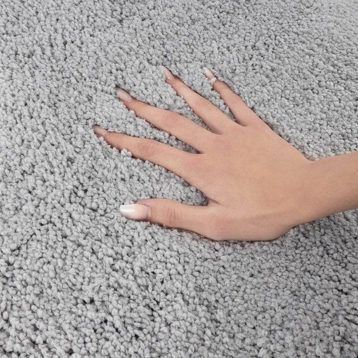 Eine Hand mit manikürten Nägeln ruht anmutig auf einem üppigen, hellgrauen Teppich und unterstreicht dessen weiche Textur und gemütliche Erscheinung.