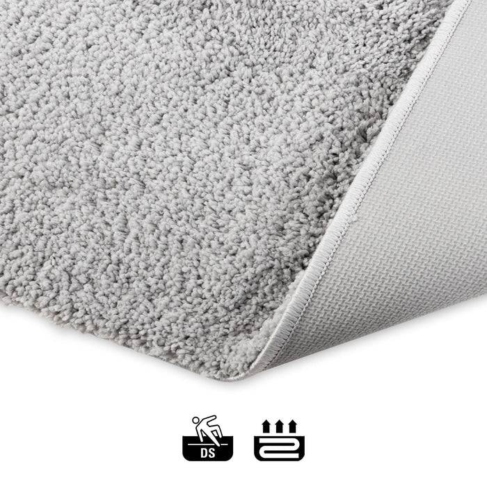 Eine Nahaufnahme eines hellgrauen Teppichs mit einer leicht umgerollten Ecke, die die Unterseite offenbart. Darunter befinden sich Wasch- und Trockensymbole für Pflegeanweisungen.