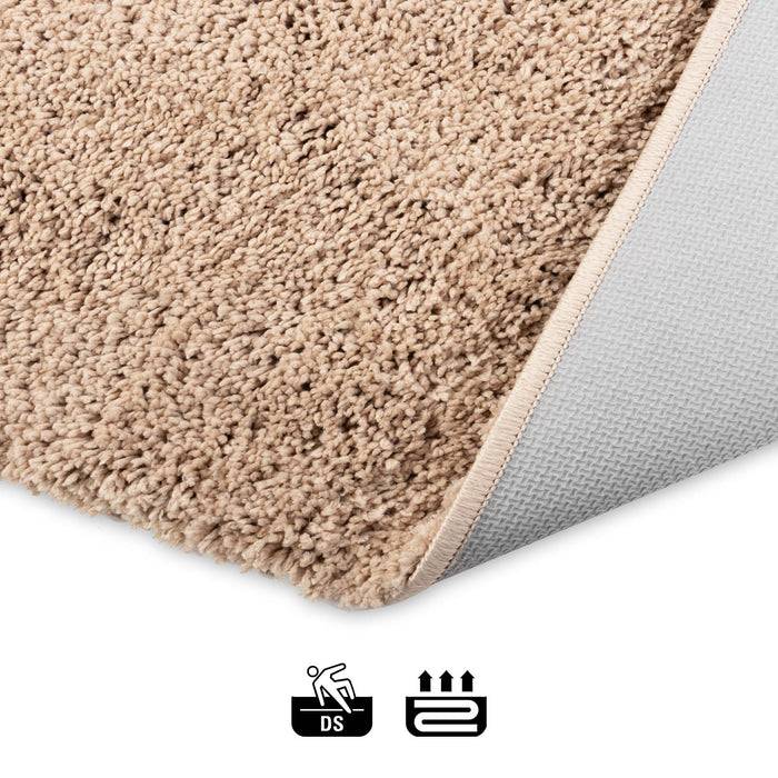 Das Bild zeigt eine Ecke eines beigen Teppichs, die umgeklappt ist und die rutschfeste Rückseite offenlegt. Darunter befinden sich Symbole, die 'DS' und Hitzebeständigkeit anzeigen.