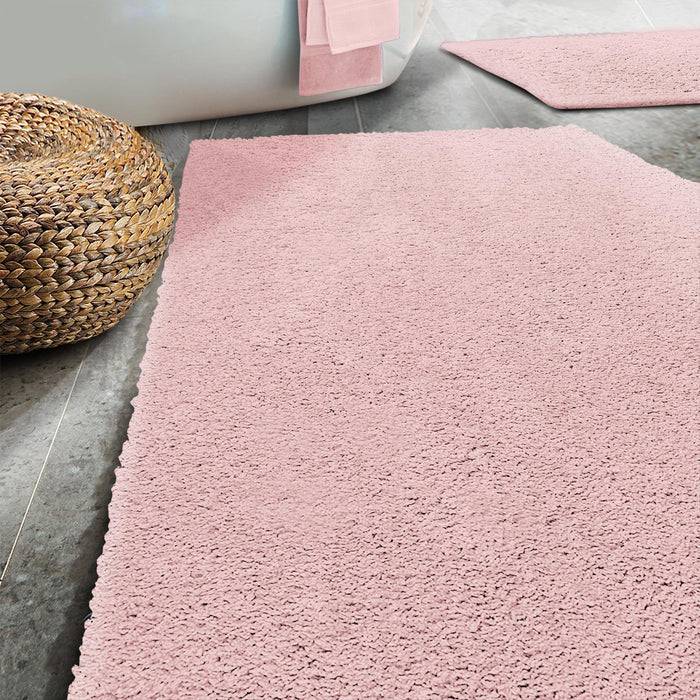 Ein modernes Badezimmer mit einer flauschigen rosa Badematte auf einem grauen Fliesenboden, mit einem geflochtenen Korb und zusammengelegten rosa Handtüchern im Hintergrund.