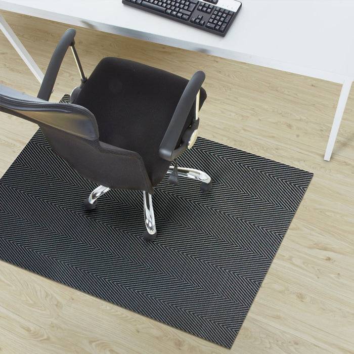 Bürostuhl auf einem schwarz-weiß gemusterten Teppich, unter einem weißen Schreibtisch mit sichtbarer Tastatur auf der Oberseite, auf einem Holzboden platziert.