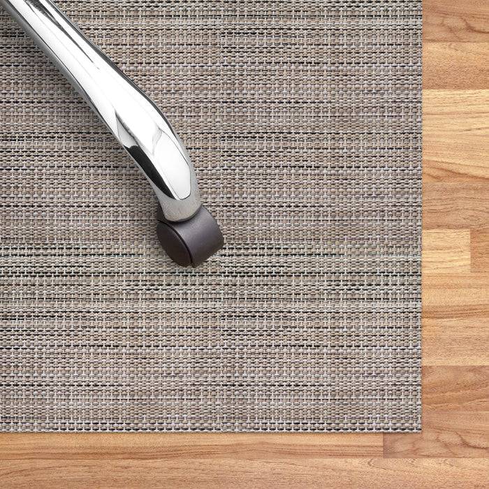 Eine Nahaufnahme eines verchromten Stuhlbeins auf einem strukturierten grauen Teppich, der auf einem Holzboden platziert ist, wobei der Kontrast zwischen den Oberflächen hervorgehoben wird.