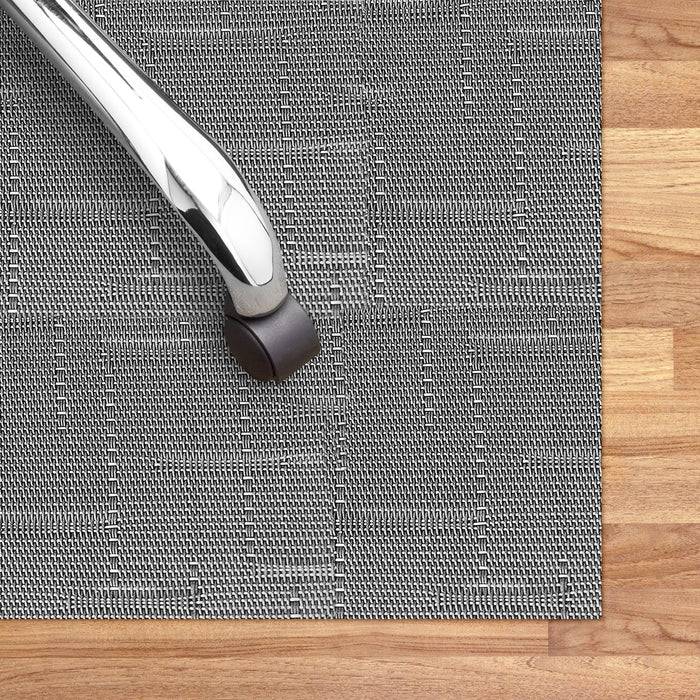 Ein verchromtes Stuhlbein auf einer strukturierten grauen Stoffmatte, die auf einem Holzboden platziert ist.