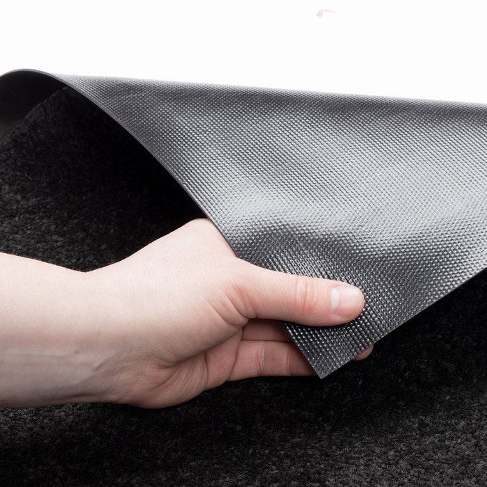 Eine Hand hebt eine Ecke einer strukturierten, rutschfesten schwarzen Matte auf einer weißen Oberfläche an und zeigt die Flexibilität der Matte und ihre Unterseite.