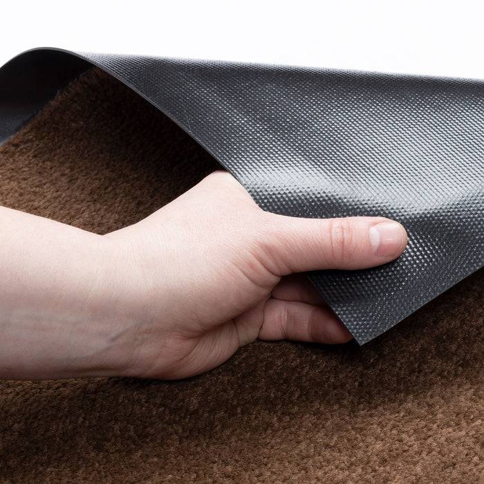 Eine Hand, die die Kante einer dunkelgefärbten Matte anhebt, zeigt ihre strukturierte Oberfläche und das darunter liegende Material über einem braunen Teppich.