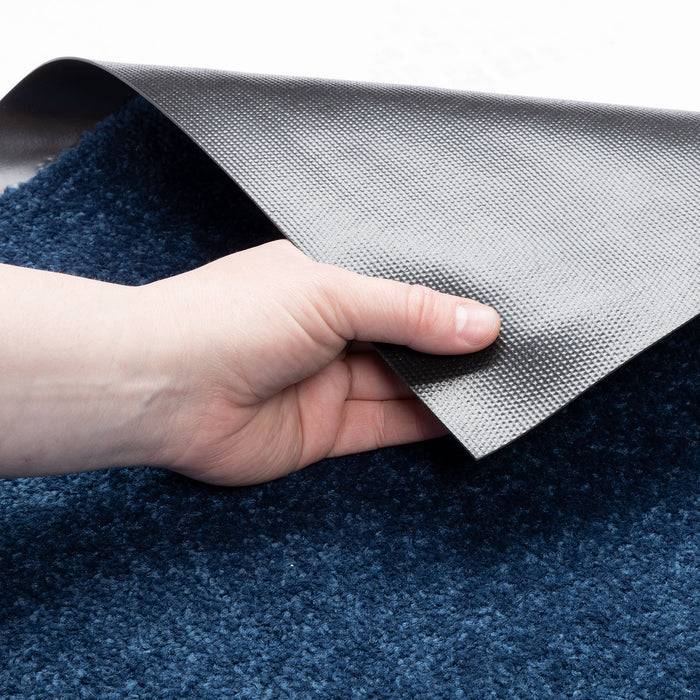 Eine Hand hebt die Ecke eines strukturierten, rutschfesten Teppichs auf einem blauen Teppich an und zeigt die Unterseite des Teppichs mit einer grauen, gepunkteten Oberfläche.