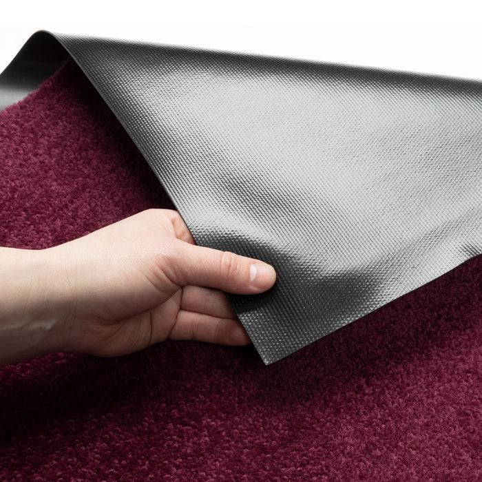 Hand hält graue Rückseite eines dunkelroten Teppichs, wobei der Texturkontrast zwischen glatter Rückseite und weichem Flor gezeigt wird.
