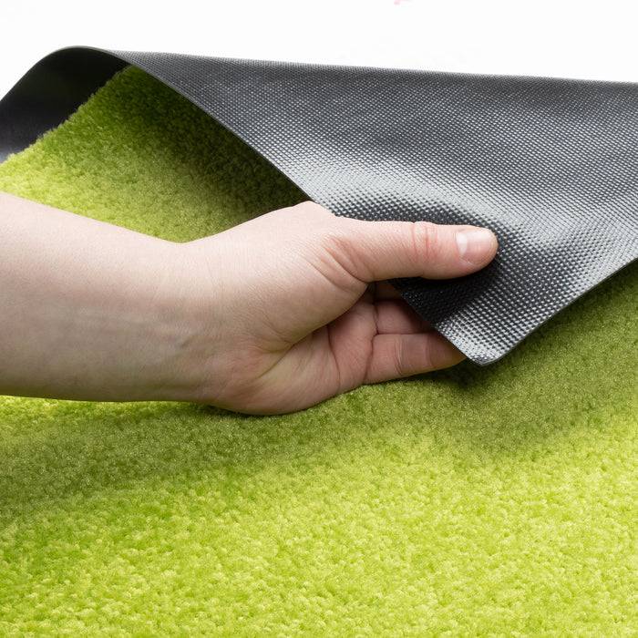 Eine Hand hebt eine Ecke eines leuchtend grünen Teppichs, wobei dessen schwarze Unterseite sichtbar wird und Textur sowie Farbkontrast demonstriert werden.