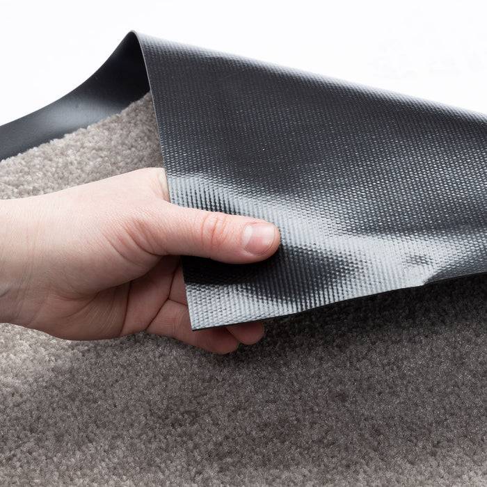 Eine Hand hebt die Ecke eines grauen Teppichs an, um seine schwarze rutschfeste Unterseite zu zeigen, was das Material des Teppichs und sein Sicherheitsmerkmal demonstriert.