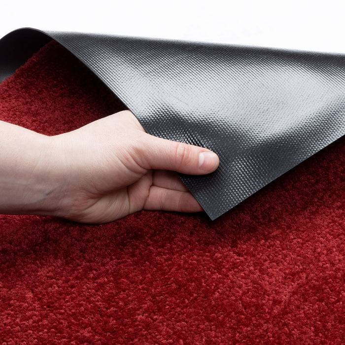 Eine Hand hebt die Ecke eines roten Teppichs an und enthüllt eine strukturierte schwarze rutschfeste Unterseite, die seine Antirutsch-Eigenschaften auf Bodenbelägen veranschaulicht.