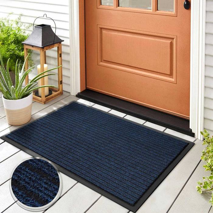 Eine dunkelblaue Fußmatte liegt vor einer geschlossenen Holztür, begleitet von einer Pflanze und einer Laterne auf einer Veranda. Eine Detailaufnahme zeigt die Textur der Matte.