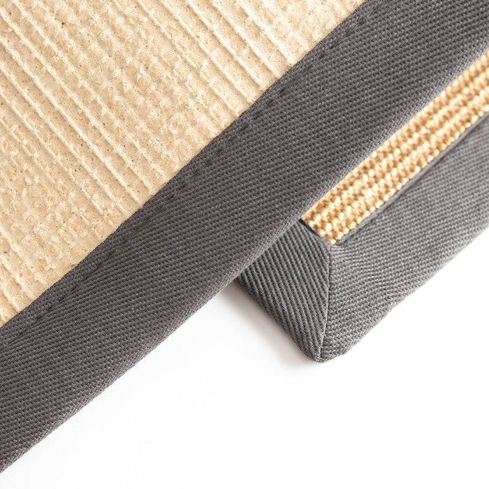 Nahaufnahme eines beigen strukturierten Stoffes mit grauer Borte, möglicherweise eine Ecke eines Kissens oder einer Matte, die Details der Stoffwebung und Nähte zeigt.