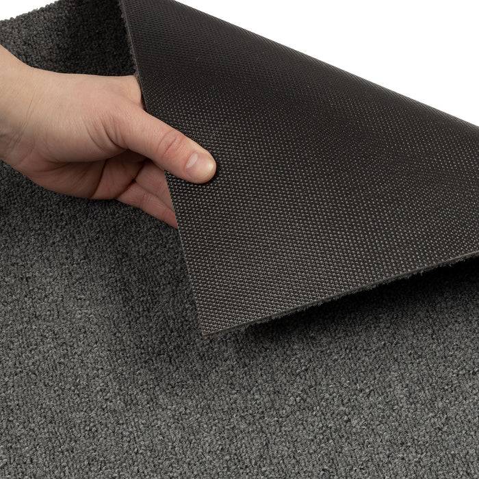 Eine Hand hebt eine dunkle, strukturierte Rückseite unter einem grauen Teppich an, um rutschfeste Eigenschaften zu zeigen und sichere Platzierung auf Böden zu betonen.